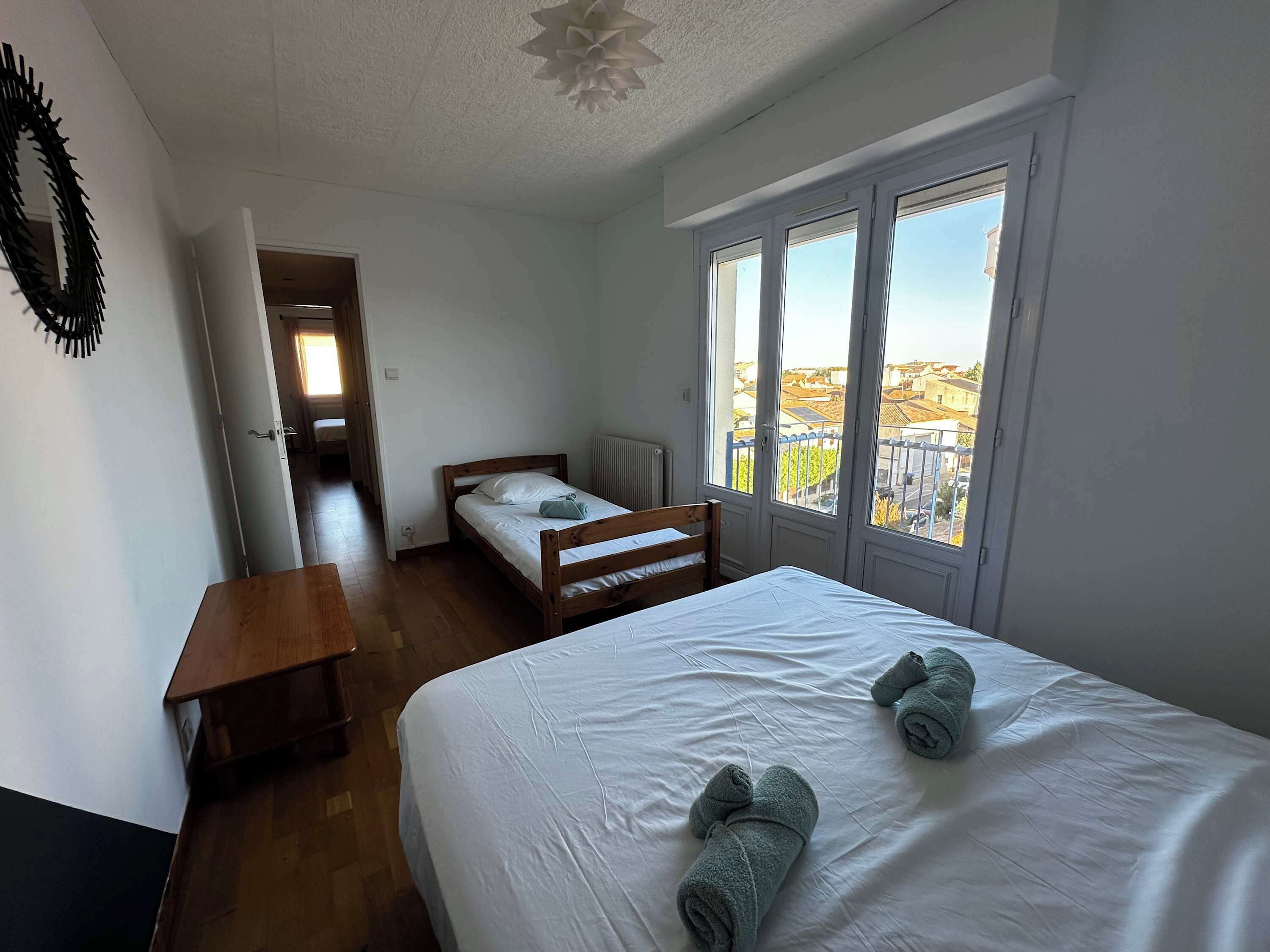 Une chambre avec un grand lit, une table de chevet en bois, une fenêtre donnant sur des immeubles et une porte menant à une autre pièce.