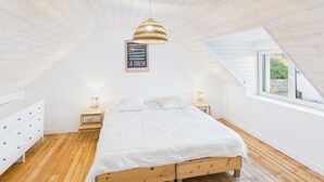 A bedroom with a sloped ceiling, a wooden bed, two bedside tables with lamps, a white dresser, and a window with a view of greenery.