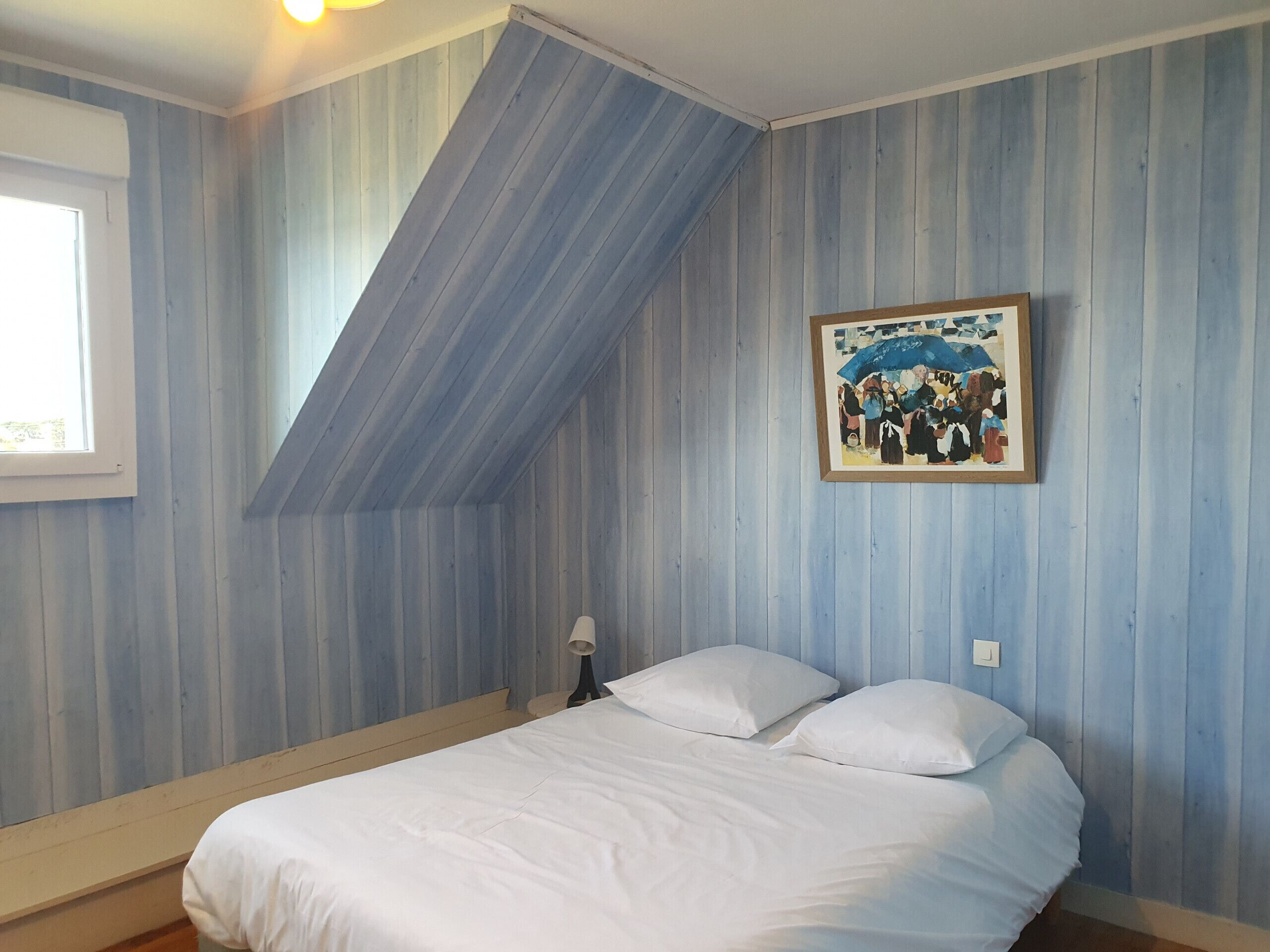 A bedroom with a sloped ceiling, a bed with white bedding, a framed picture on the wall, and a window with blinds.