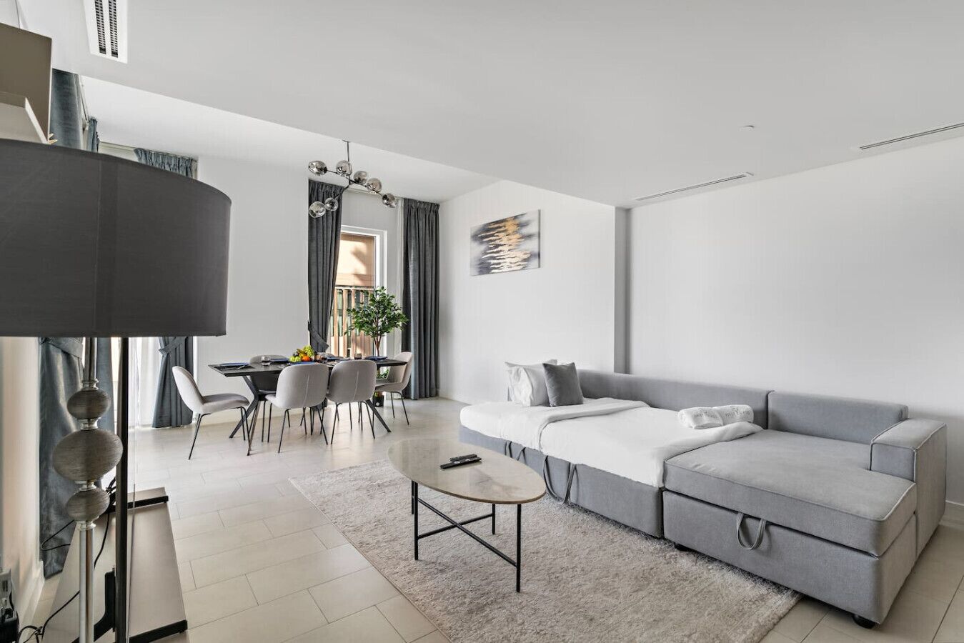A modern living room with a grey sofa, a dining table with chairs, and a large window with curtains.