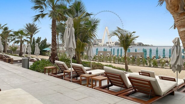 A poolside area with lounge chairs and umbrellas, a palm tree, and a view of a water body and a Ferris wheel in the background. - Spacious Dubai Marina 2BR | Great Views, Top Location (JBR)