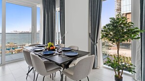A dining area with a table set for four, large windows with curtains, a view of the sea and buildings, and a potted plant. - Spacious Dubai Marina 2BR | Great Views, Top Location (JBR)