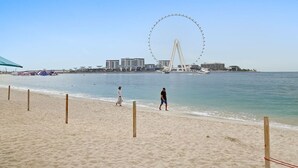 A beach with a Ferris wheel in the background, sandy shore, and a few people walking. - Spacious Dubai Marina 2BR | Great Views, Top Location (JBR)
