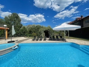 Una piscina con sillas de descanso y sombrillas, rodeada de árboles y una casa.