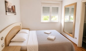 A neatly made bed with pillows and folded towels, a window with blinds, and a mirror on the wall. - Holiday Home Nadalina (Vrgorac)