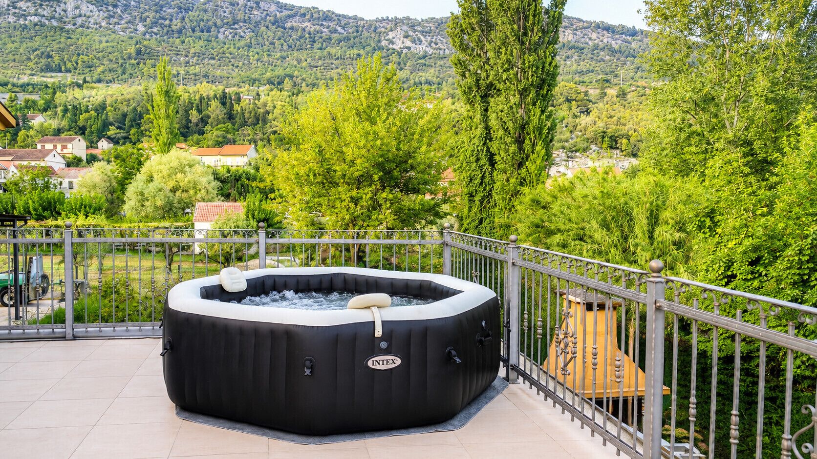 A hot tub on a balcony with a metal railing, overlooking a green landscape with trees and houses.