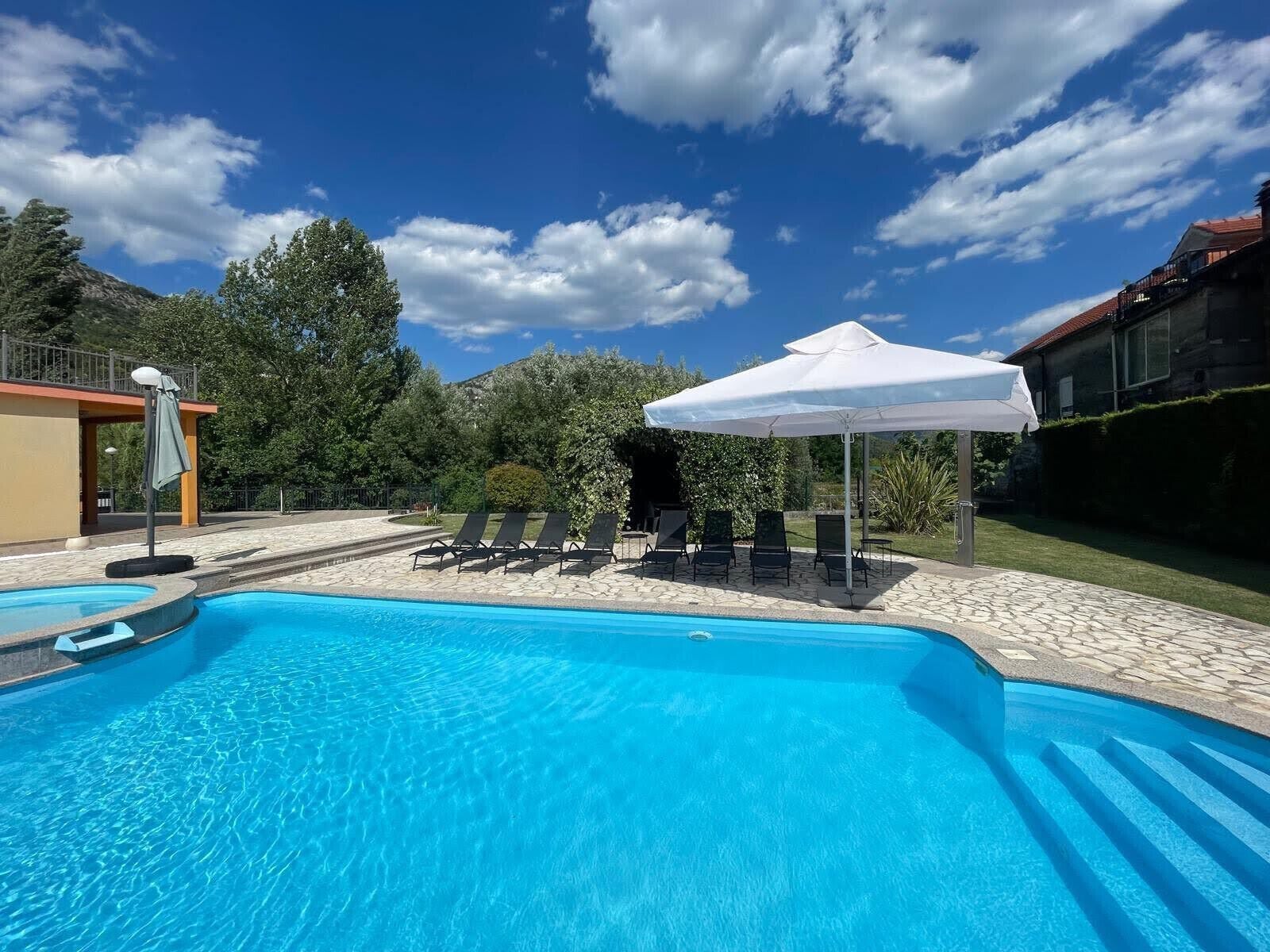 Une piscine entourée d’arbres et d’un bâtiment, avec des chaises longues et des parasols.