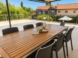 Een houten eettafel met stoelen, een glazen schaal en een kaars op een terras met uitzicht op een huis en groen.