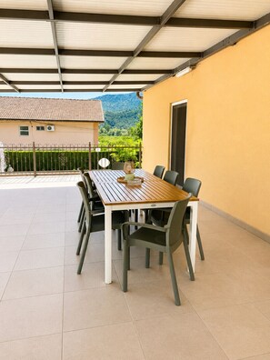 Une terrasse couverte avec une table et des chaises, un mur jaune et une vue sur une maison et des montagnes.