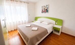 Una cama bien tendida con un cabecero verde, una mesita de noche con cajón y una obra de arte colgada en la pared.
