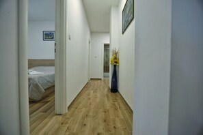 A narrow hallway with wooden flooring, a bed with a white duvet, and a vase with yellow flowers on the right wall. - Holiday Home Amaya (Vrgorac)
