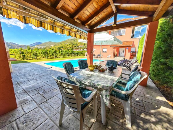 A covered outdoor seating area with a pool, chairs, and a table with bowls of fruit. - Holiday Home Amaya (Vrgorac)