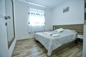 A bedroom with a wooden floor, a bed with a grey bedspread, a nightstand, and a window with floral curtains.