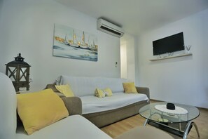 A living room with a sofa, a glass coffee table, a TV mounted on the wall, and a painting of sailboats.