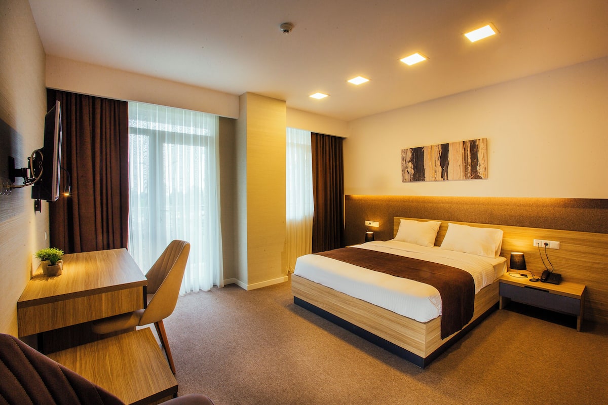 A hotel room with a bed, desk, chair, and a small table with a plant.