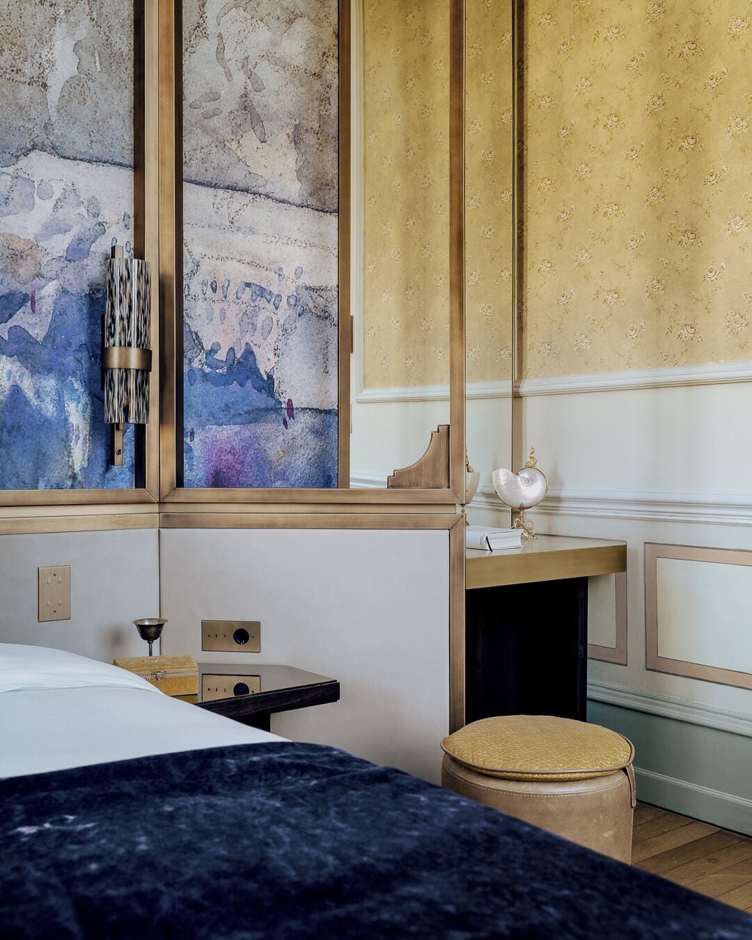 A hotel room with a bed, a small table, a decorative lamp, and a wall with a floral pattern.