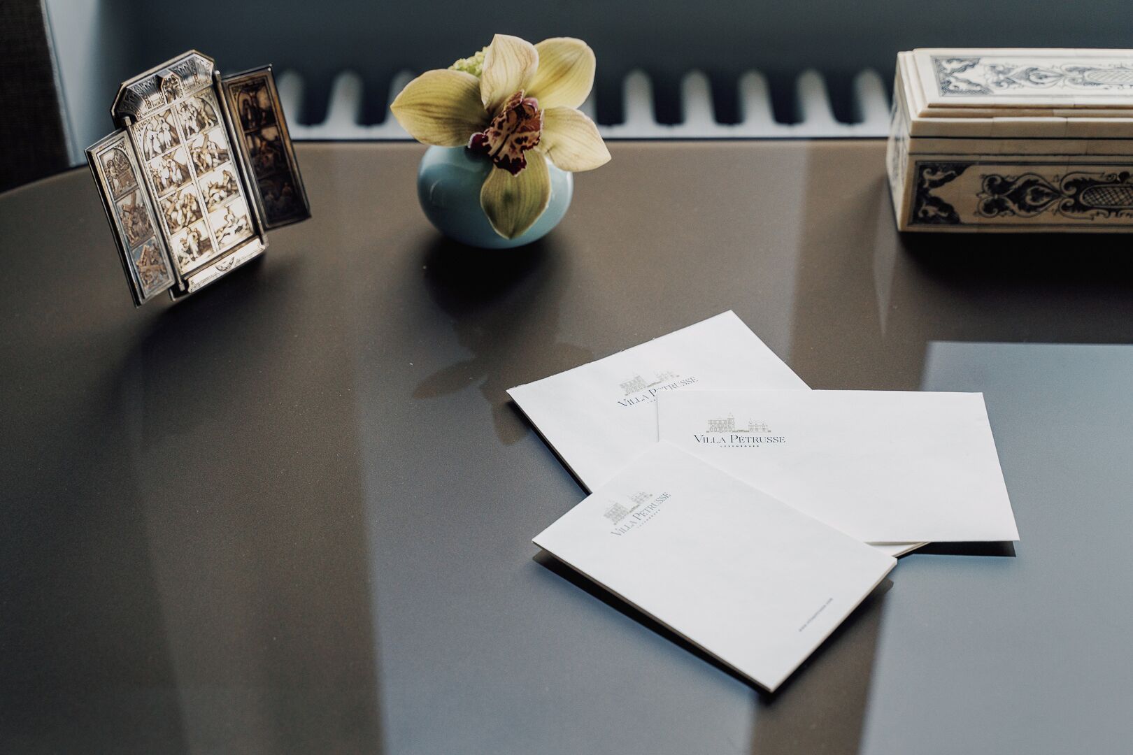 A table with a blue vase containing a single orchid, two white envelopes with 'Villa Petruše' printed on them, and a silver photo frame with multiple images.