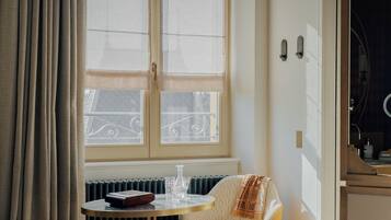A hotel room with a bed, a round table with a chair, a radiator, and a window with curtains.