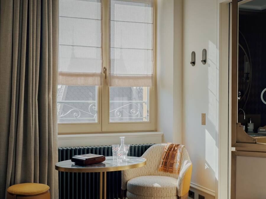 A hotel room with a bed, a round table with a chair, a radiator, and a window with curtains.