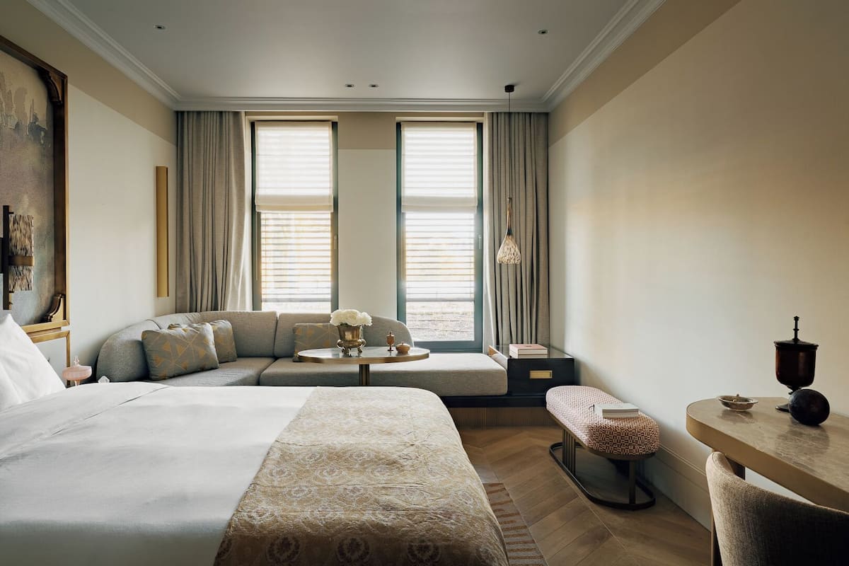 Habitación de hotel con una cama grande, un sofá, una pequeña mesa y un banco. Hay dos ventanas con cortinas, un cuadro en la pared y una lámpara colgante.