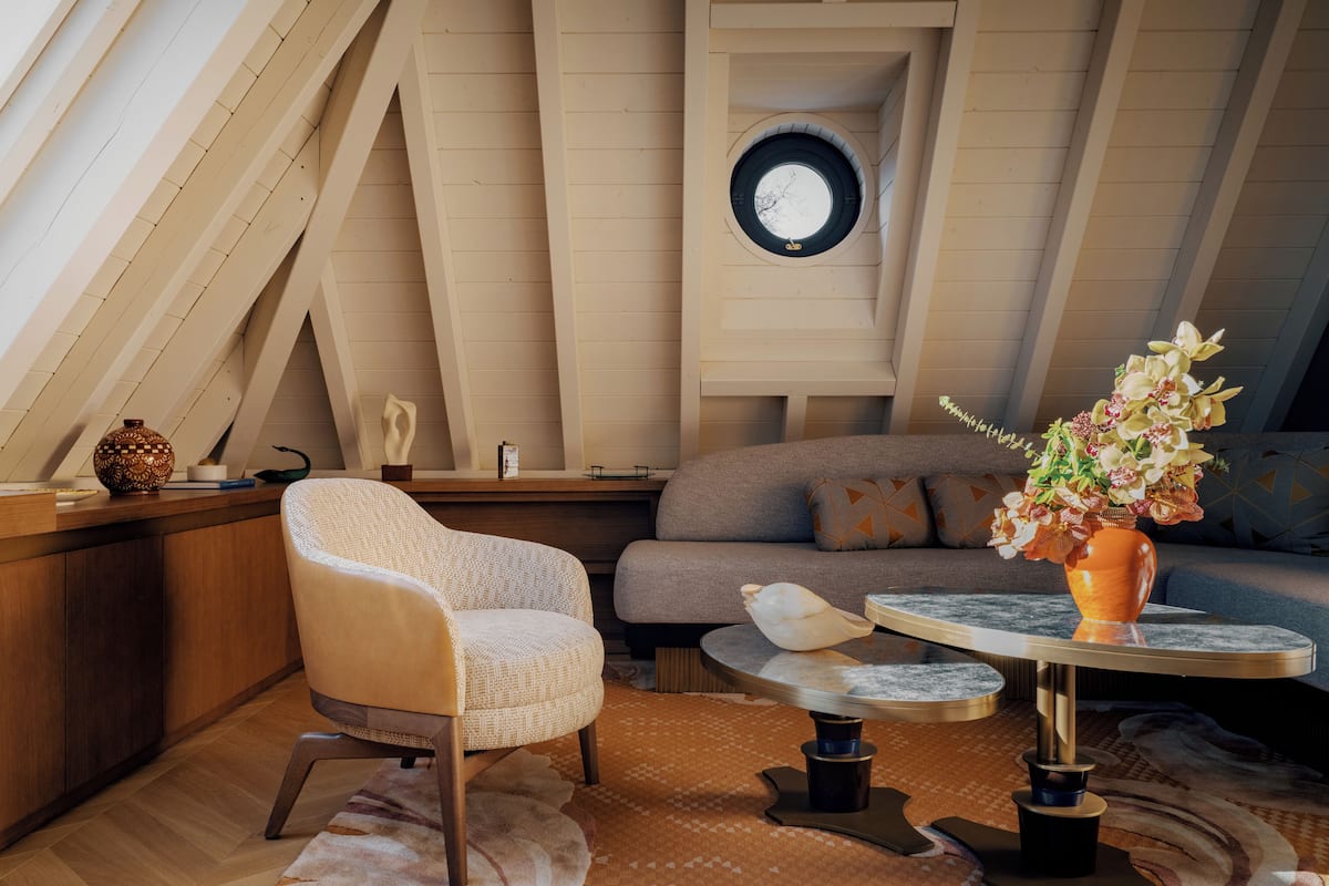 Un dormitorio tipo loft acogedor, con techo inclinado, tragaluz circular, un sofá cómodo, una silla, una mesita redonda y un jarrón con flores.