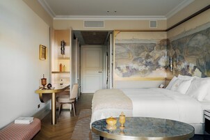 A hotel room with a large bed, a round table, a desk with a chair, and a painting on the wall.