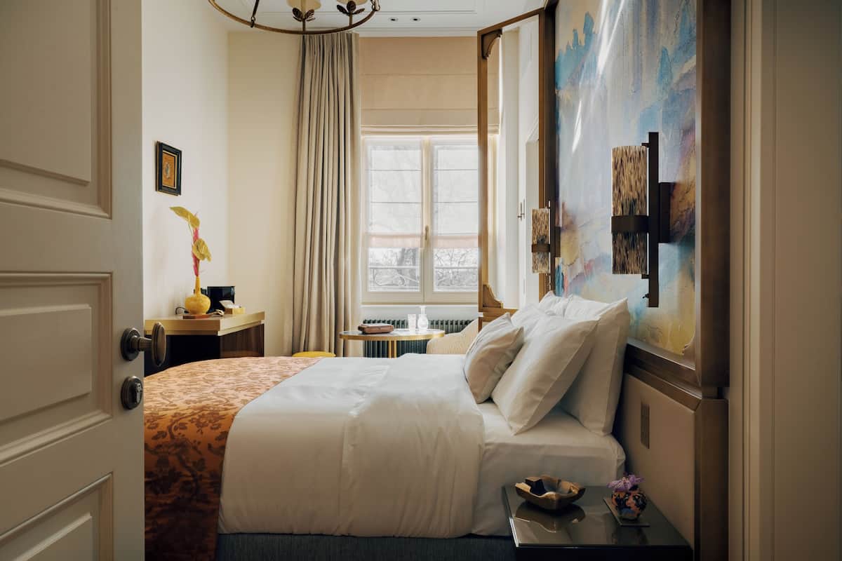 Habitación de hotel con cama, mesita de noche, ventana con vistas y un cuadro en la pared.
