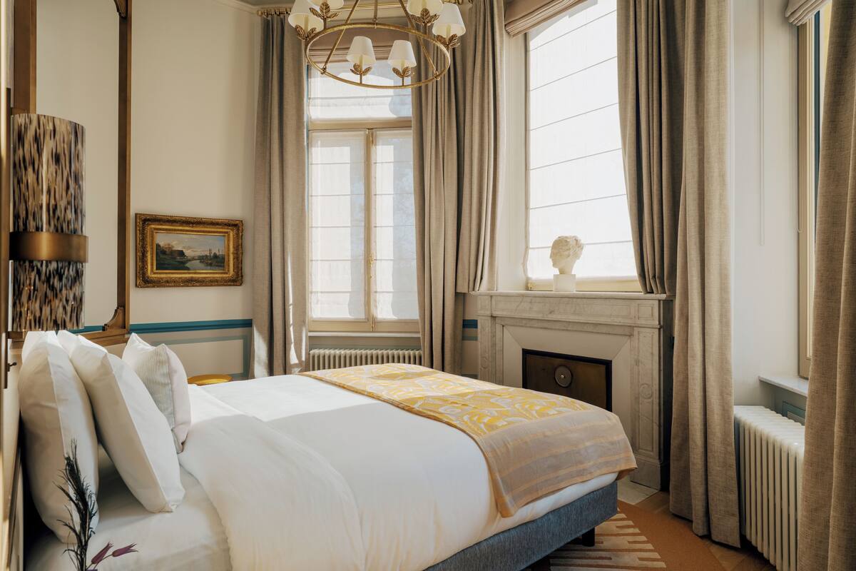 Habitación de hotel con una cama grande, chimenea, candelabro y vistas al exterior por la ventana.