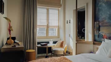 Quarto de hotel com cama, escrivaninha com um vaso, cadeira, radiador e uma janela grande com cortinas.