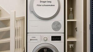 A compact, white laundry room with a front-loading washing machine and dryer, a small shelf with cleaning supplies, and a white railing. - 20 minutes to Amsterdam (Almere)