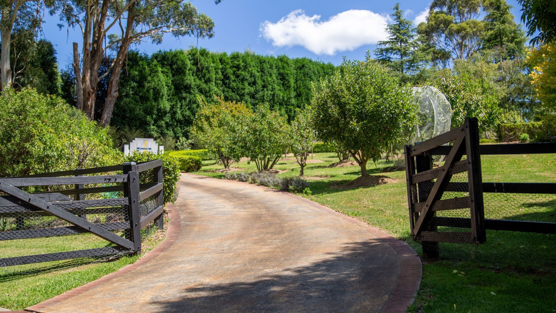A paved pathway leading through a landscaped area with a wooden gate, surrounded by trees and greenery.