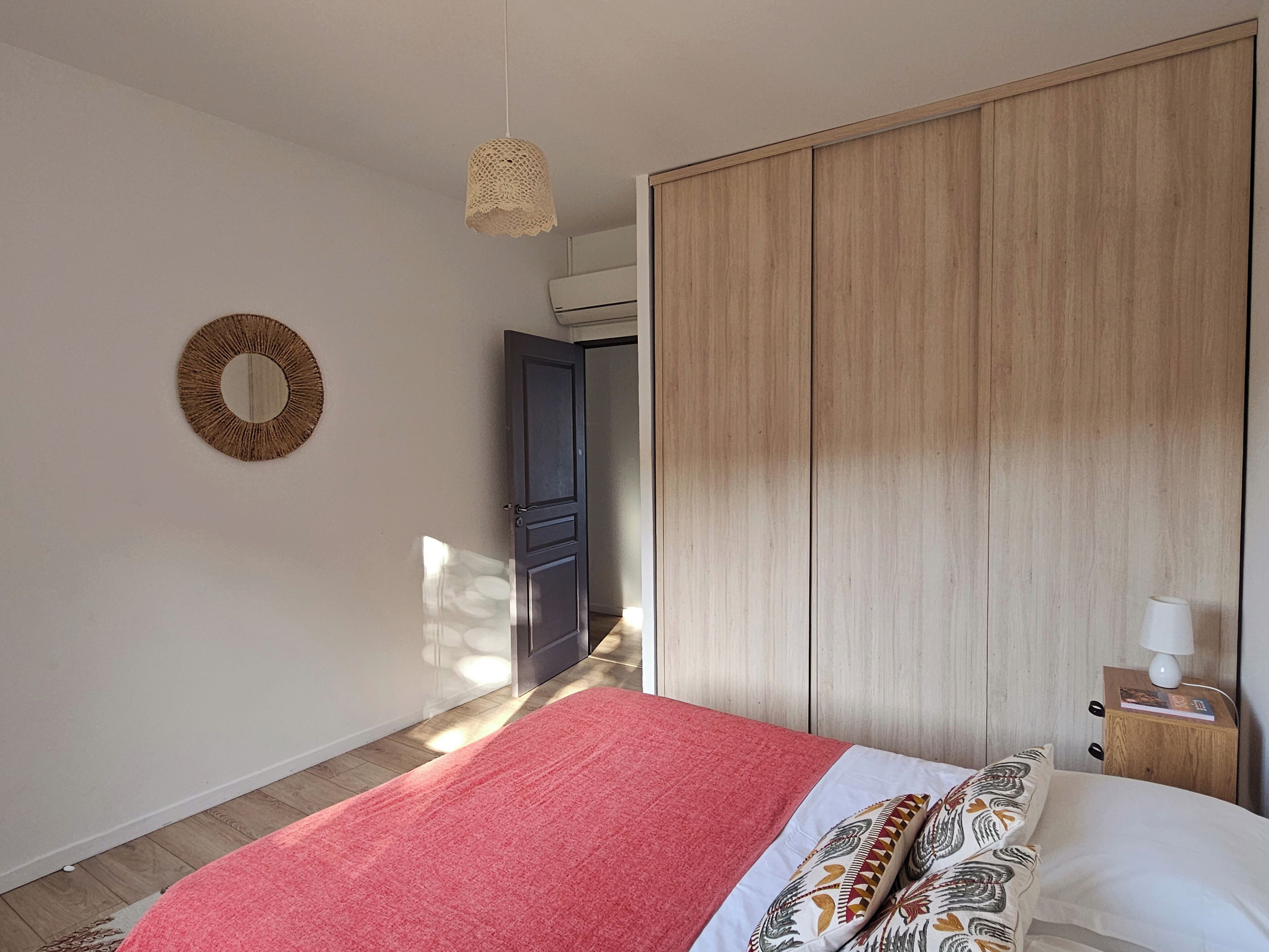 Une chambre à coucher avec un lit, une armoire en bois, un miroir rond et une petite table de chevet avec une lampe.