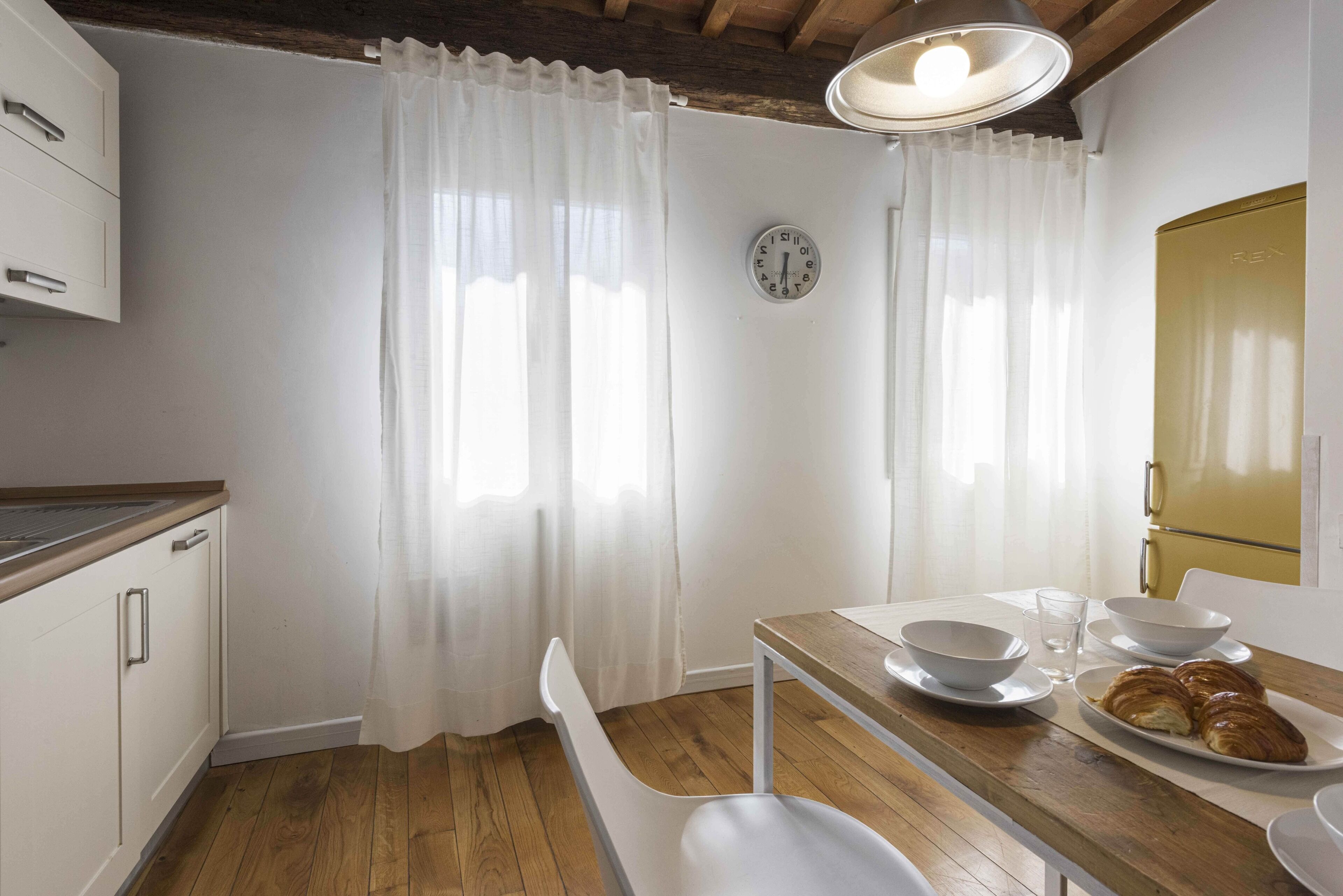 A kitchen with white cabinets, a wooden countertop, a dining table with chairs, a clock, and a window with white curtains.