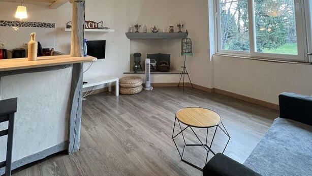 A spacious living area with a wooden ceiling, a large window offering a view of trees, a central seating area with a round table, and a kitchen counter with various items. - Le petit gite dans la prairie (Revel)