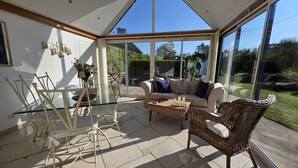 A sunlit conservatory with a glass ceiling, a sofa, wicker chairs, and a glass dining table.