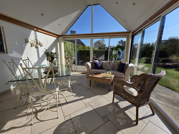 A sunlit conservatory with a glass ceiling, a sofa, wicker chairs, and a glass dining table.