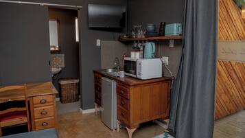 Cocina compacta con microondas, tostadora y refrigerador pequeño. Hay un mueble de madera con cajones y una cesta de mimbre. Un televisor de pantalla plana está montado en la pared sobre la encimera.