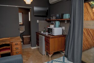 Cocina compacta con microondas, tostadora y refrigerador pequeño. Hay un mueble de madera con cajones y una cesta de mimbre. Un televisor de pantalla plana está montado en la pared sobre la encimera.