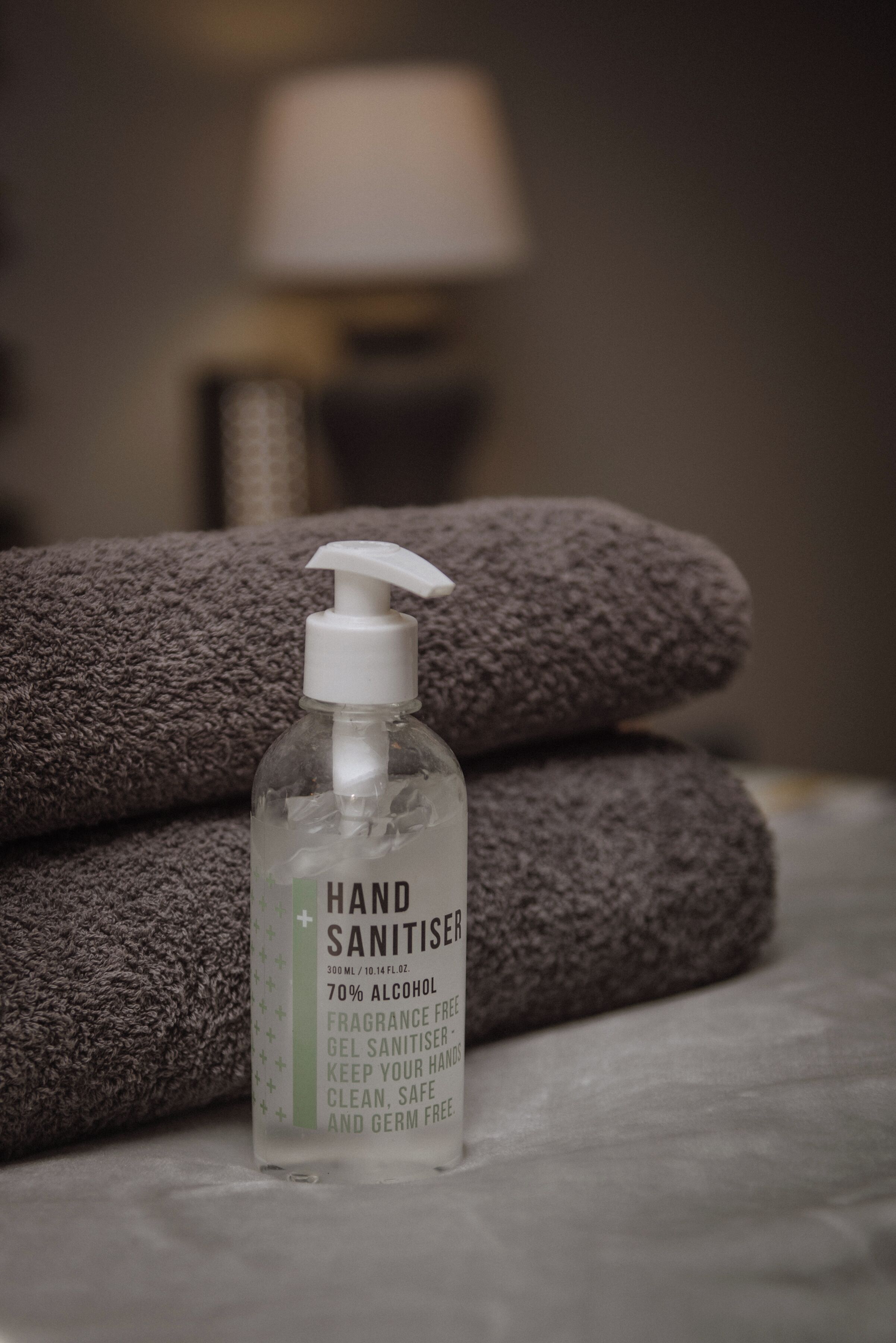 A hand sanitizer bottle labeled "HAND SANITISER" with 70% alcohol, placed in front of folded towels on a bed.