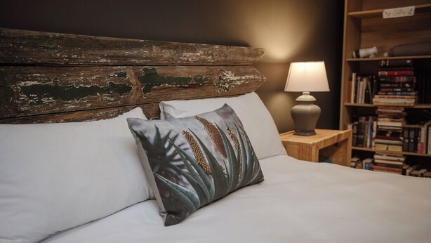 Un letto con lenzuola bianche, una testiera in legno e un cuscino decorato con un motivo naturalistico.