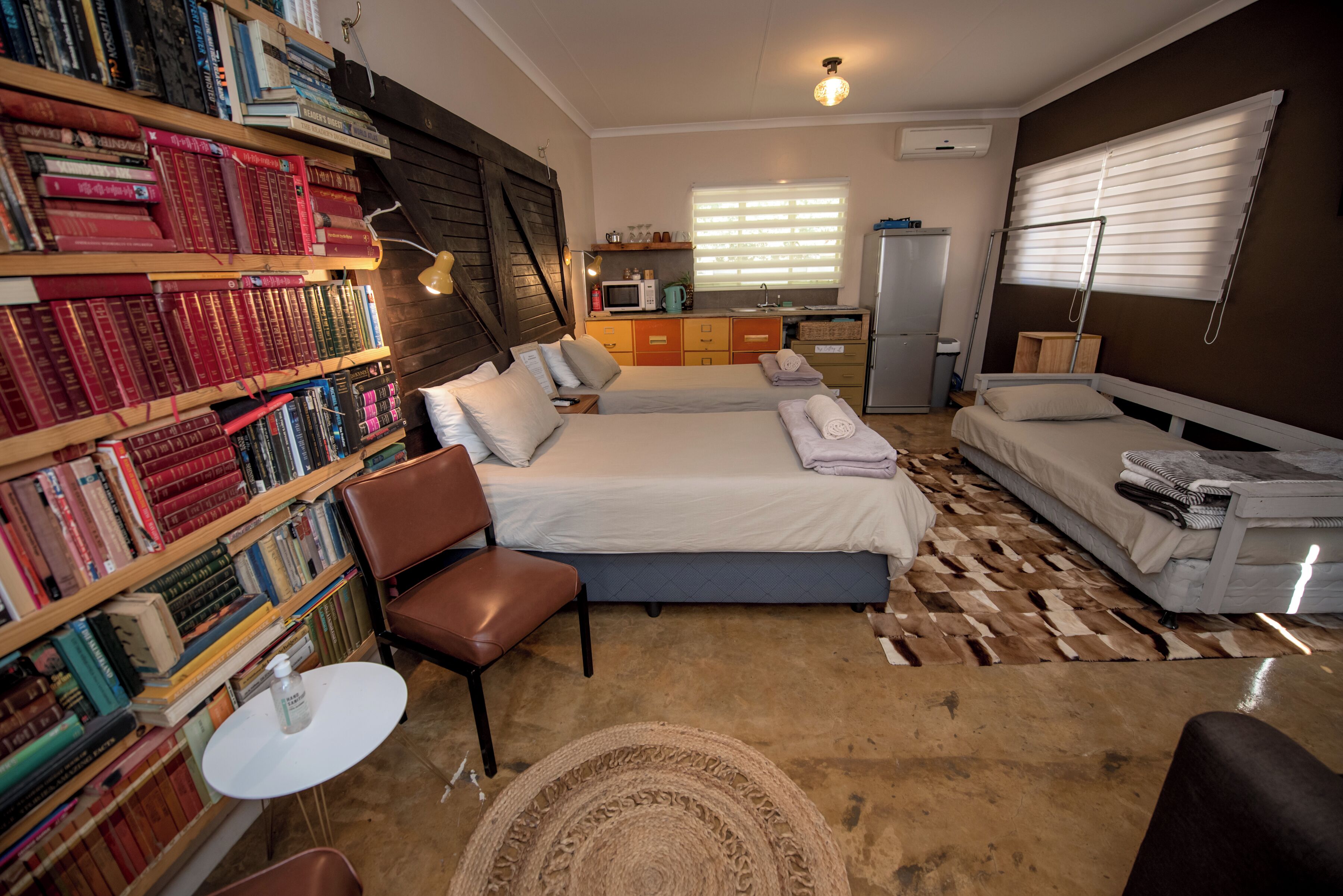 A compact hotel room with a large bookshelf, a bed, a desk, and a kitchenette.