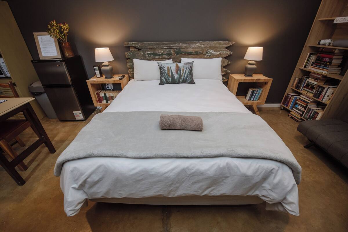 Ein ordentlich bezogenes Bett mit grauer Decke, zwei Nachttischlampen, einem Holzkopfstück und einem Bücherregal auf der rechten Seite.