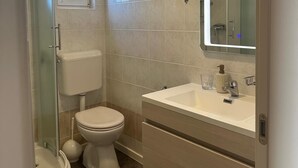 A bathroom with a glass-enclosed shower, a toilet, a sink with a mirror, and a window. - Apartment Matkovic 4 (Jadranovo)