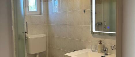 Un baño con cabina de ducha de vidrio, inodoro, lavamanos con espejo y ventana.