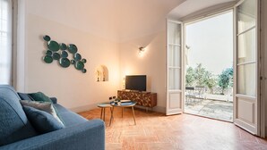 Ein Wohnzimmer mit einer Couch, einem Fernseher auf einer Holzkommode, einem Wandbild und Blick ins Freie durch eine offene Tür.