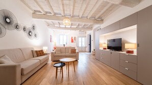 Un grand salon comprenant un canapé, deux petites tables, un téléviseur et un plafond avec des poutres apparentes.