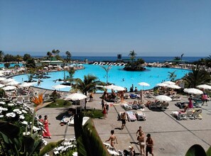 A large outdoor swimming pool surrounded by lounge chairs and umbrellas, with palm trees and a clear view of the ocean. - Stunning 3 Bedroom Apartment in Radazul With Ocean Views (El Rosario)
