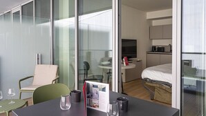 Una moderna camera d'hotel con parete in vetro, angolo ristoro con tavolo e sedie, un letto, una TV e un angolo cottura.