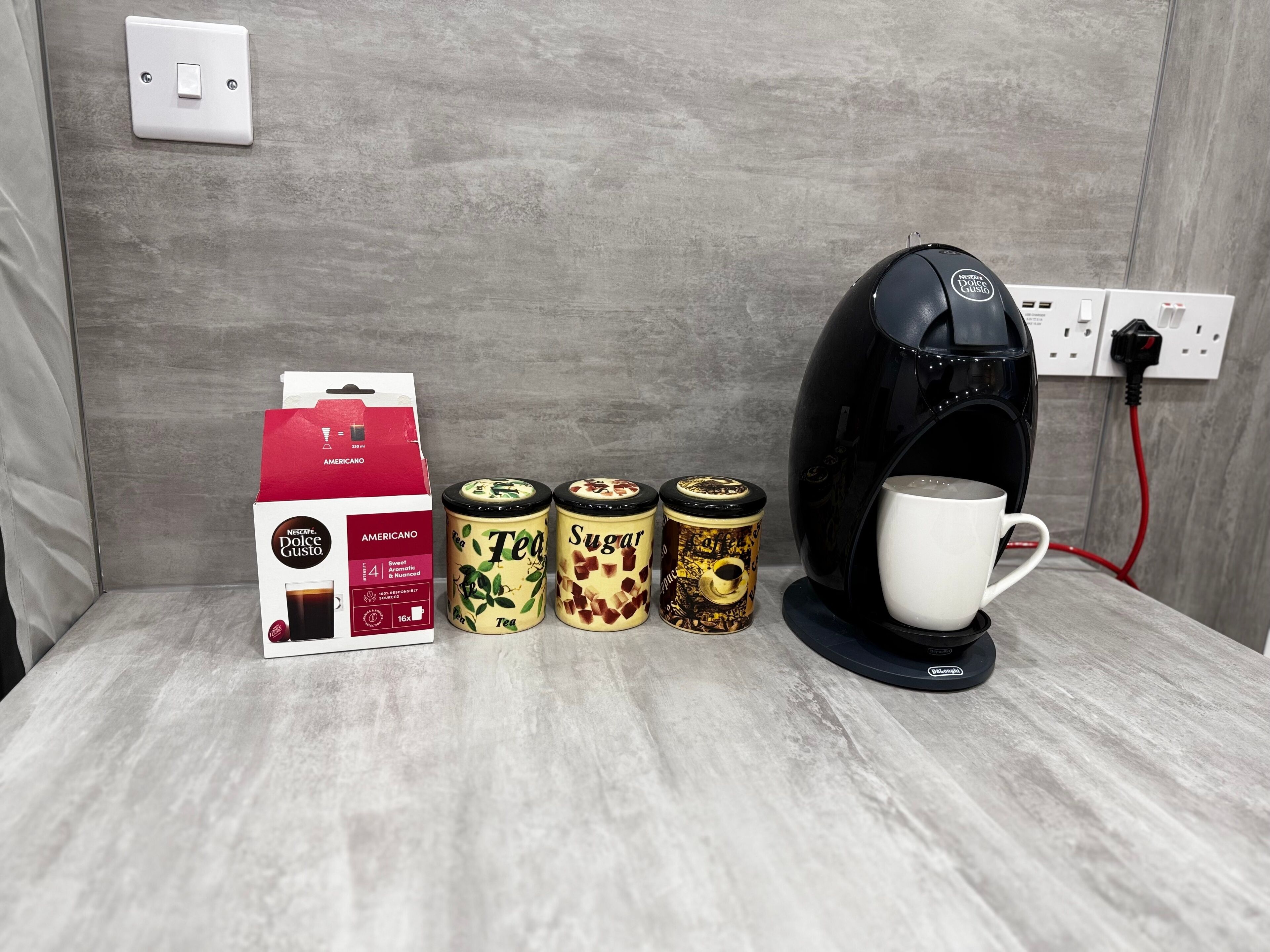 A coffee machine, a box of Nescafe Dolce Gusto Americano, and three containers of tea and sugar.