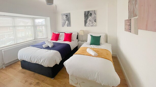 A hotel room with two beds, one with a blue bedspread and the other with a yellow one. There are two paintings on the wall, a white radiator, and a window with blinds. - BetaHouse -3BR -Pool Table -Free Parking -Spacious (Greater Manchester)